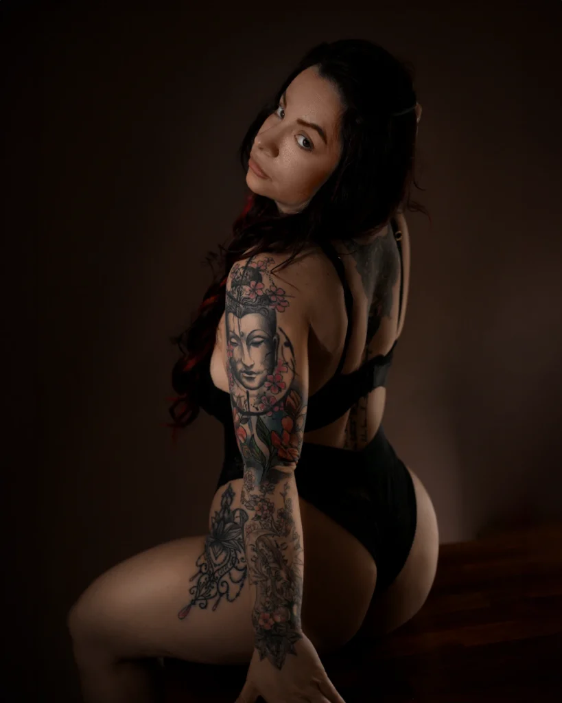 Boudoir Fotografie in Bamberg – tätowierte Frau in schwarzen Dessous sitzt auf Holzboden und blickt über die Schulter, warmes Licht betont Haut und Tattoo-Details.